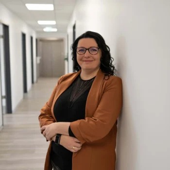 Heike Anstötz, verantwortlich für QM und Controlling, lehnt lächelnd an einer Wand in einem modernen Büroflur.
