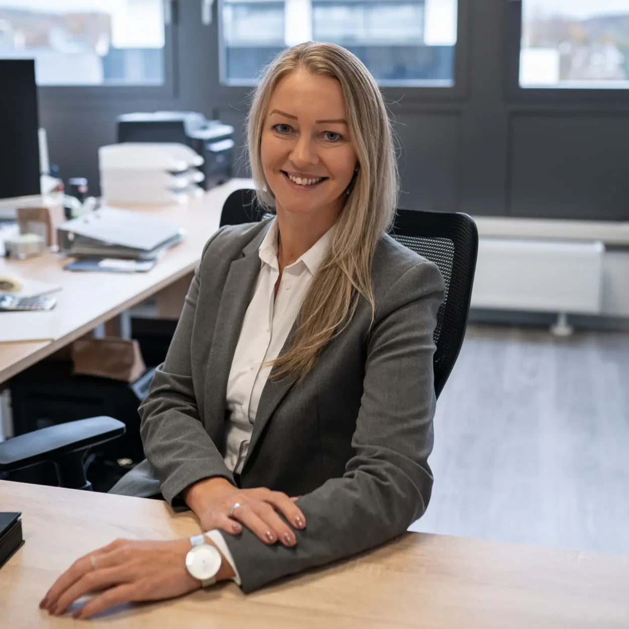 Irene Haar, Steuerberaterin und Partnerin, sitzt lächelnd an einem Schreibtisch in einem modernen Büro, mit einem professionellen grauen Blazer und weißem Hemd.