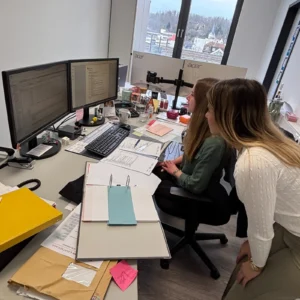 Zwei Frauen in einem Büro, die gemeinsam an einem Schreibtisch mit mehreren Monitoren, Dokumenten und Büroartikeln arbeiten, mit Blick auf einen sonnigen Ausblick durch die Fenster.