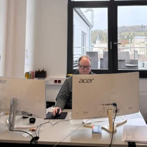 Mann in einem Büro, der konzentriert an zwei Monitoren arbeitet. Große Fenster im Hintergrund bieten einen Blick auf umliegende Gebäude.