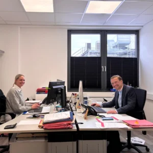 Zwei Personen in einem Büro, die an ihren Schreibtischen arbeiten und in die Kamera lächeln. Im Hintergrund ein Fenster mit Blick auf technische Anlagen und Gebäude.