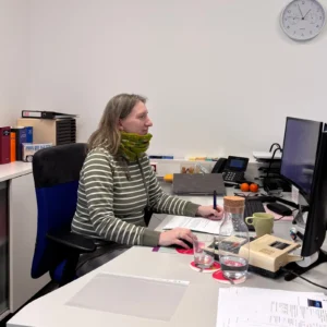 Frau in einem Büro, die an einem Computer arbeitet, umgeben von Dokumenten, Büromaterialien und einer Wanduhr im Hintergrund.