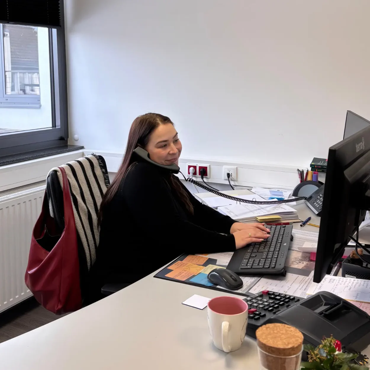 Eine Mitarbeiterin telefoniert am Schreibtisch und arbeitet am Computer. Auf dem Schreibtisch befinden sich eine Tasse, eine Tastatur, Dokumente und Büromaterialien. Im Hintergrund ist ein Fenster mit Tageslicht zu sehen.