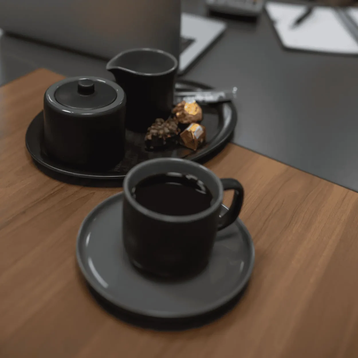 Eine schwarze Kaffeetasse auf einer Untertasse steht auf einem Holztisch, daneben eine schwarze Zuckerdose, ein Milchkännchen und einige Pralinen auf einem Tablett.