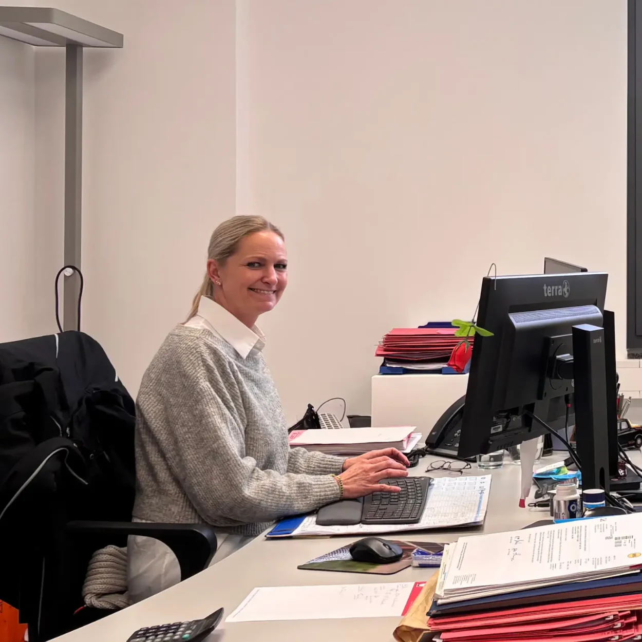 Eine Mitarbeiterin sitzt lächelnd an ihrem Schreibtisch in einem Büro, tippt auf einer Tastatur und schaut in die Kamera. Im Hintergrund sind Aktenstapel und ein Computerbildschirm sichtbar.