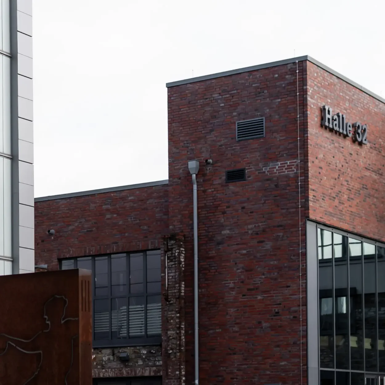 Außenansicht der Halle 32, einem modernen Backsteingebäude mit großen Fenstern, seitlich ergänzt durch eine Skulptur aus Metall.