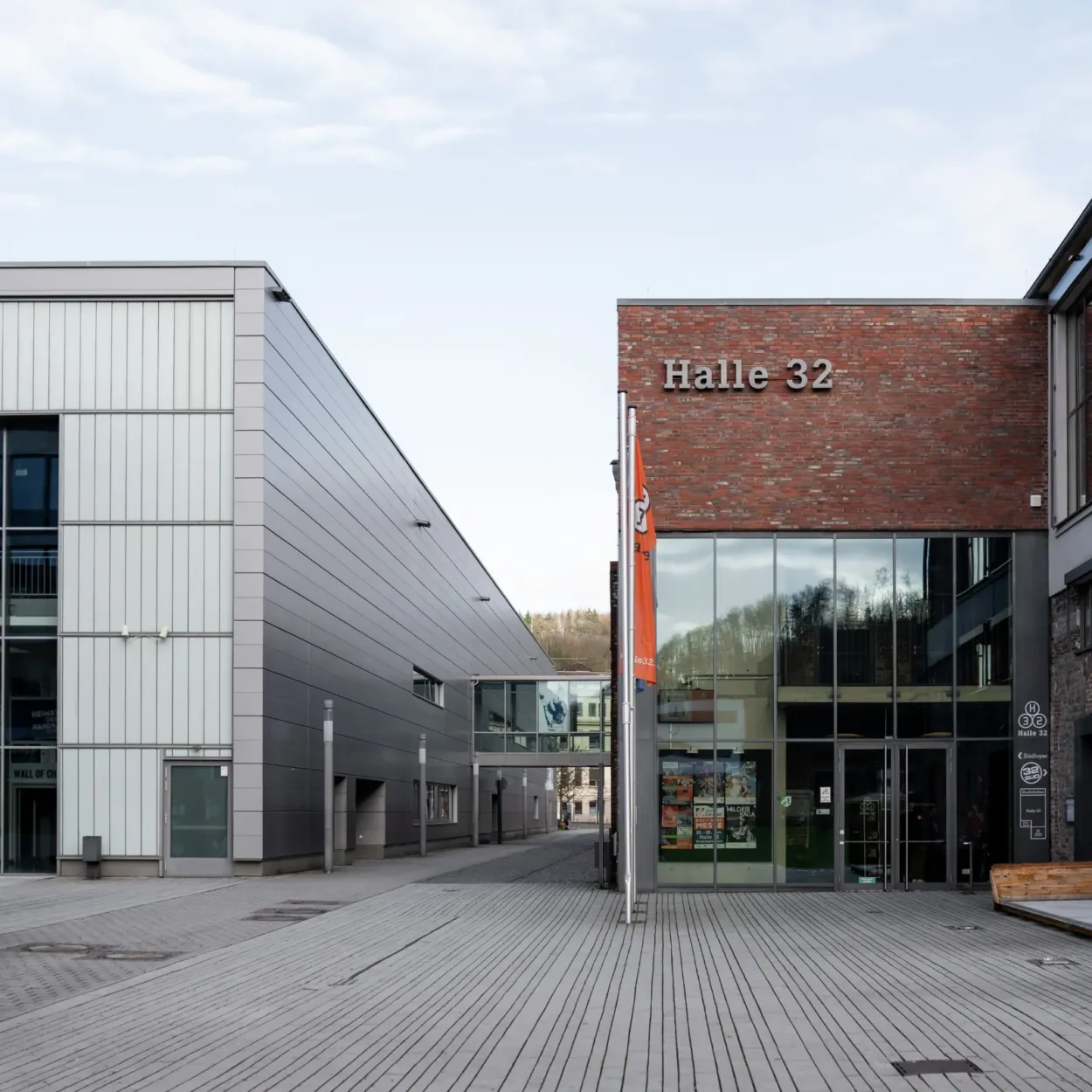 Außenansicht der Halle 32 in Gummersbach auf dem Steinmüllergelände mit moderner Architektur aus Backstein und Metall sowie einem breiten Gehweg davor.