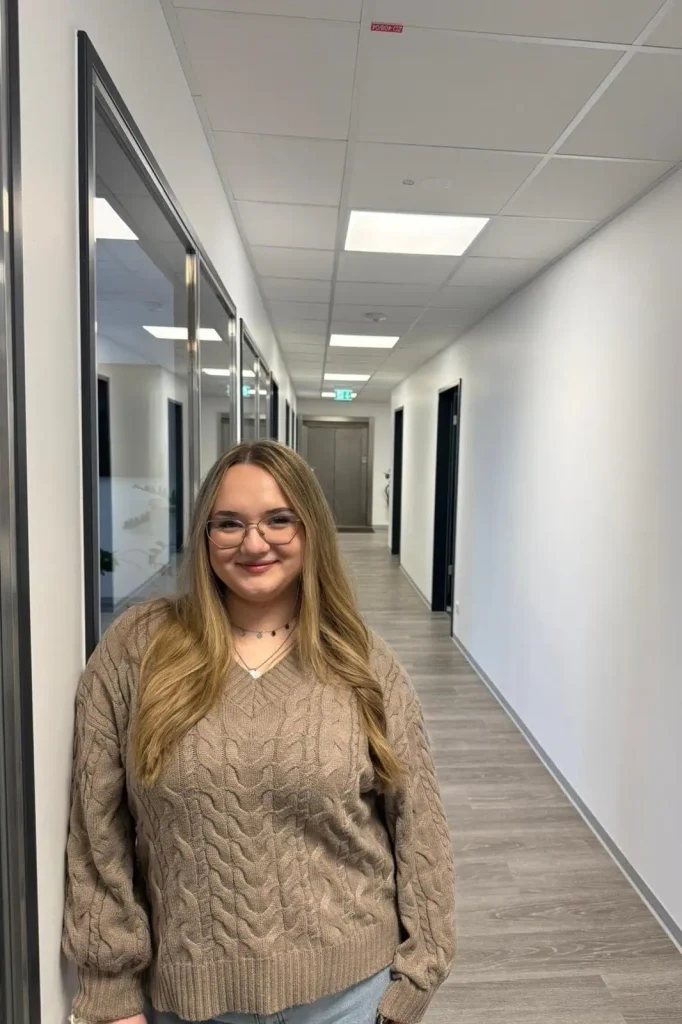 Junge Frau mit Brille und braunem Strickpullover steht lächelnd in einem modernen Büroflur.