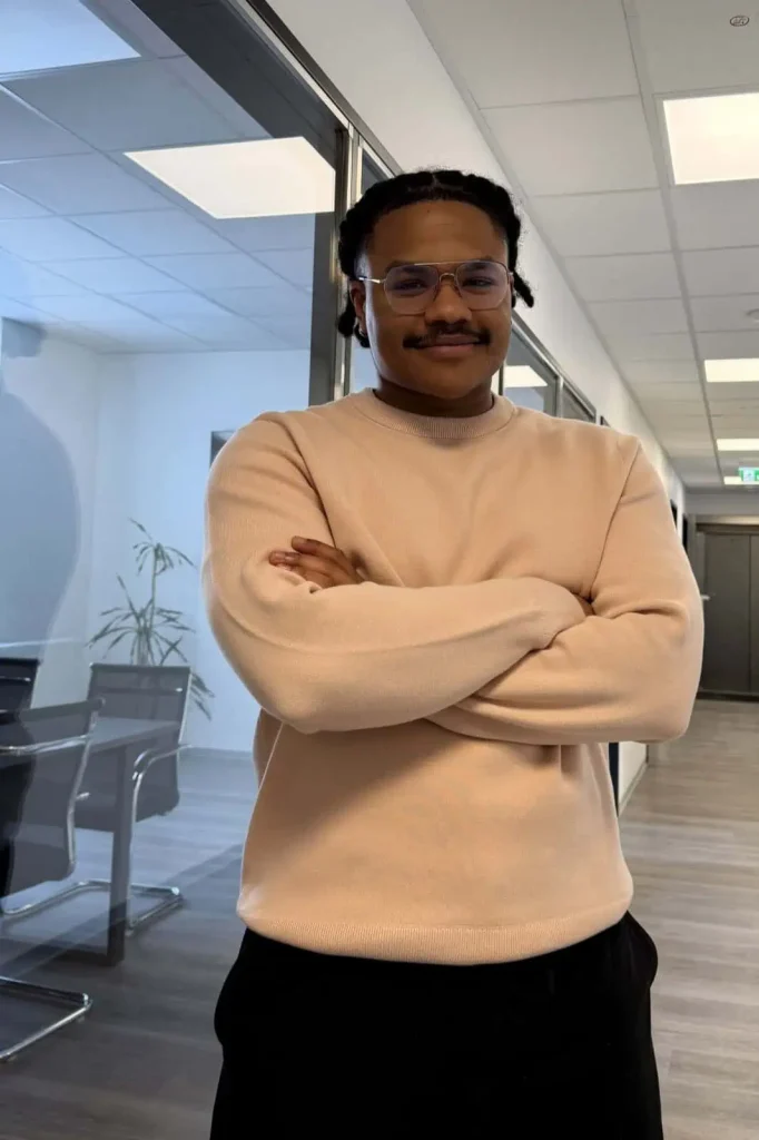 Junger Mann mit Brille und hellem Pullover steht mit verschränkten Armen in einem modernen Büroflur und lächelt.