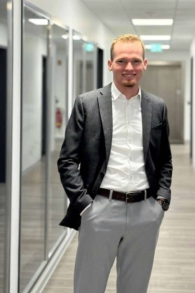 Junger Mann in weißem Hemd und dunklem Sakko steht lächelnd in einem modernen Büroflur.
