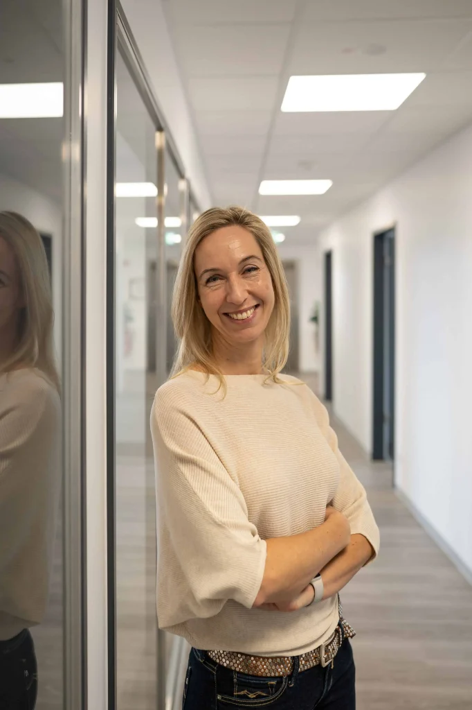 Nadja Braatz, Rechtsanwältin, steht lächelnd mit verschränkten Armen an einer Glaswand in einem modernen Büroflur.