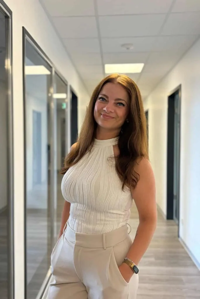 Junge Frau mit langen braunen Haaren und heller Businesskleidung steht lächelnd in einem modernen Büroflur.