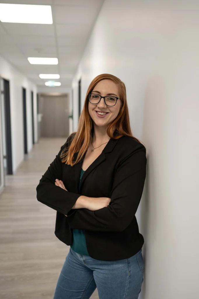 Sara-Marie Junghans, Steuerfachangestellte, lehnt lächelnd an einer Wand in einem modernen Büroflur.